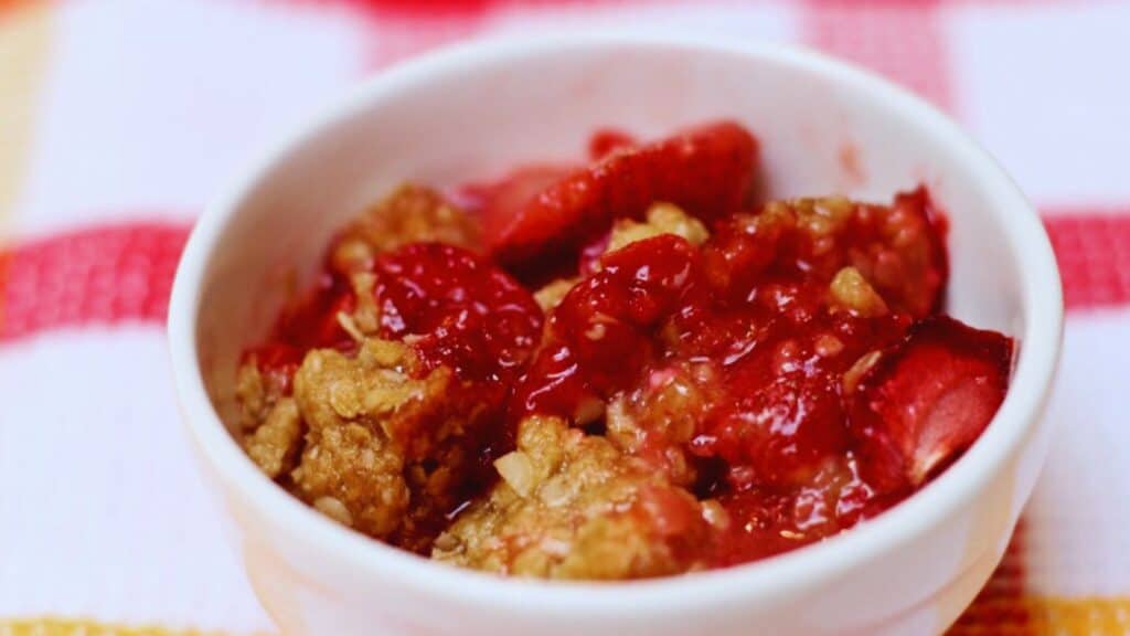 A bowl of strawberry crumble dessert on a striped tablecloth.