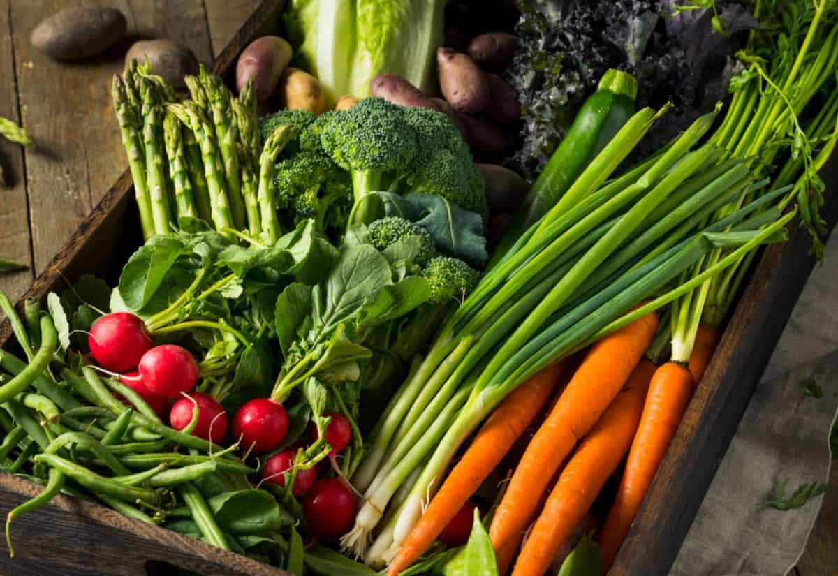 A variety of fresh spring produce, including carrots, asparagus, broccoli, radishes, and green onions, neatly arranged in a wooden crate.