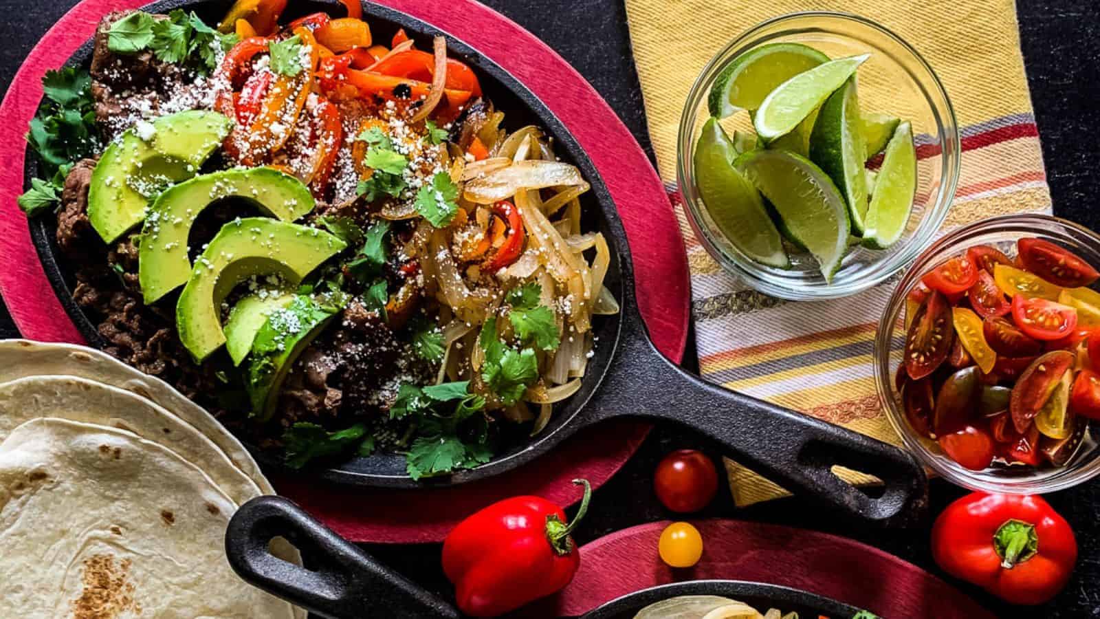 A cast iron skillet with hot beef fajitas and seared vegetables alongside bowls of lime wedges, tomatoes, and tortillas.