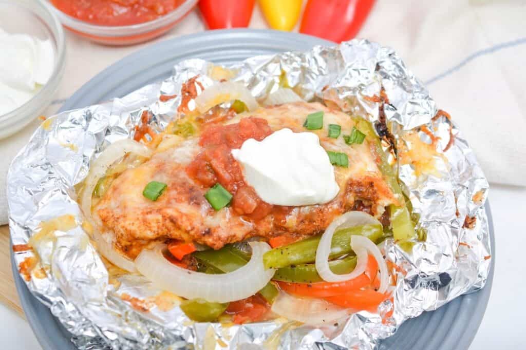 Grilled chicken breast topped with salsa, melted cheese, and a dollop of sour cream, served with peppers and onions on a foil-wrapped plate.