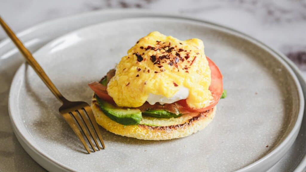 A plate with eggs benedict featuring a poached egg, hollandaise sauce, and avocado slices on an english muffin, garnished with spices, accompanied by gold cutlery. Perfect for Mother's Day recipes.