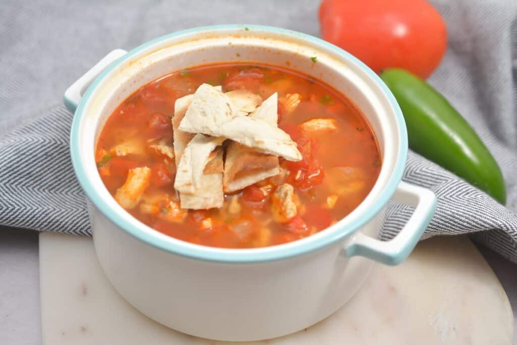 A bowl of tomato-based soup garnished with tortilla strips, with fresh vegetables in the background.