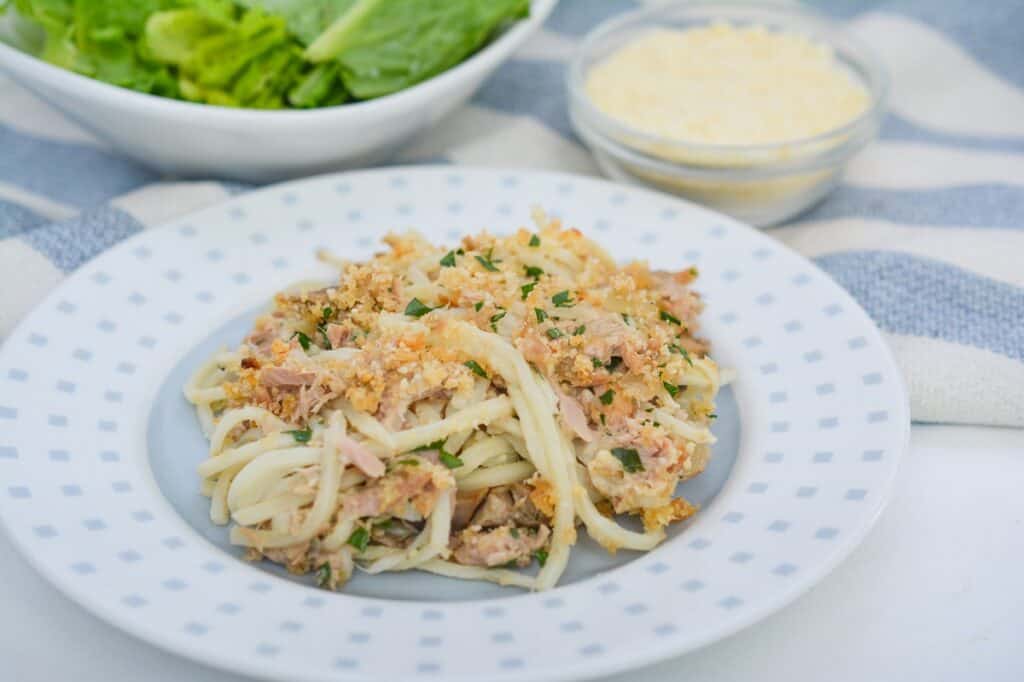 A plate of pasta with tuna, garnished with breadcrumbs and parsley, accompanied by grated cheese and a bowl of greens.