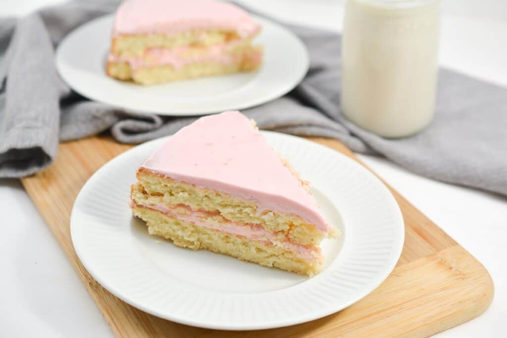 A slice of layered cake with pink frosting on a white plate, accompanied by a glass of milk.