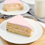 A slice of layered cake with pink frosting on a white plate, accompanied by a glass of milk.