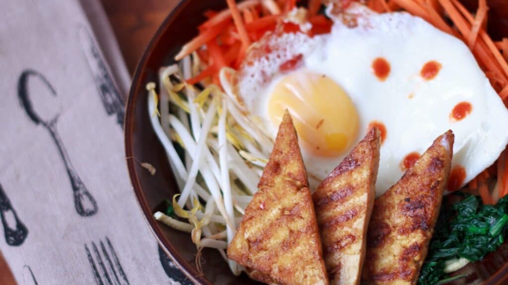 A bowl of food featuring a fried egg, stir-fried greens, shredded carrots, bean sprouts, and grilled tofu triangles.