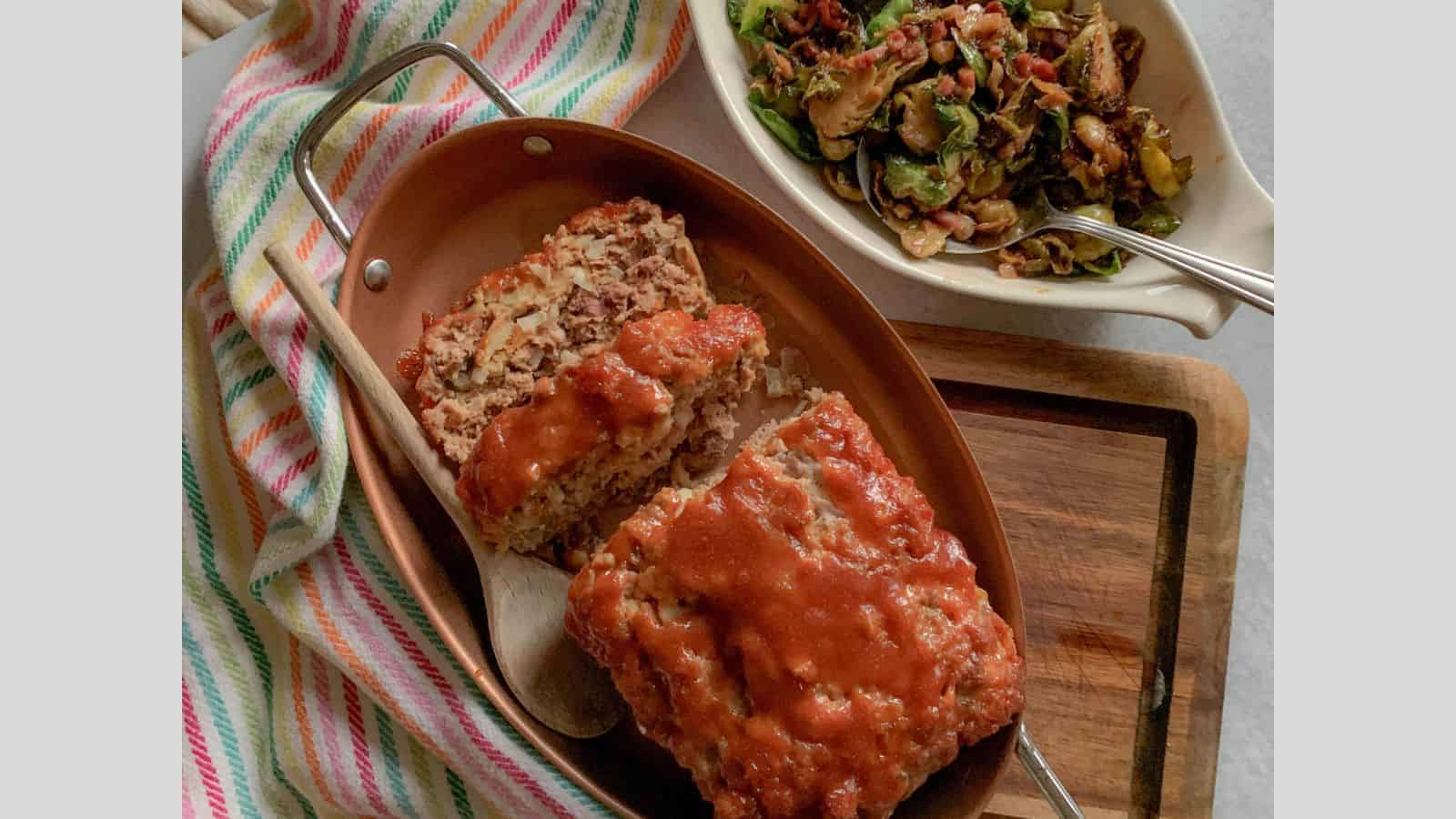 A half-sliced meatloaf in a copper baking dish with a bowl of Brussels sprouts alongside.