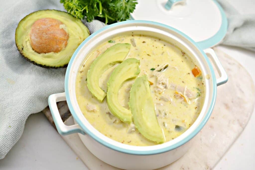 A bowl of creamy soup topped with avocado slices, served with a side of bread and a whole avocado.