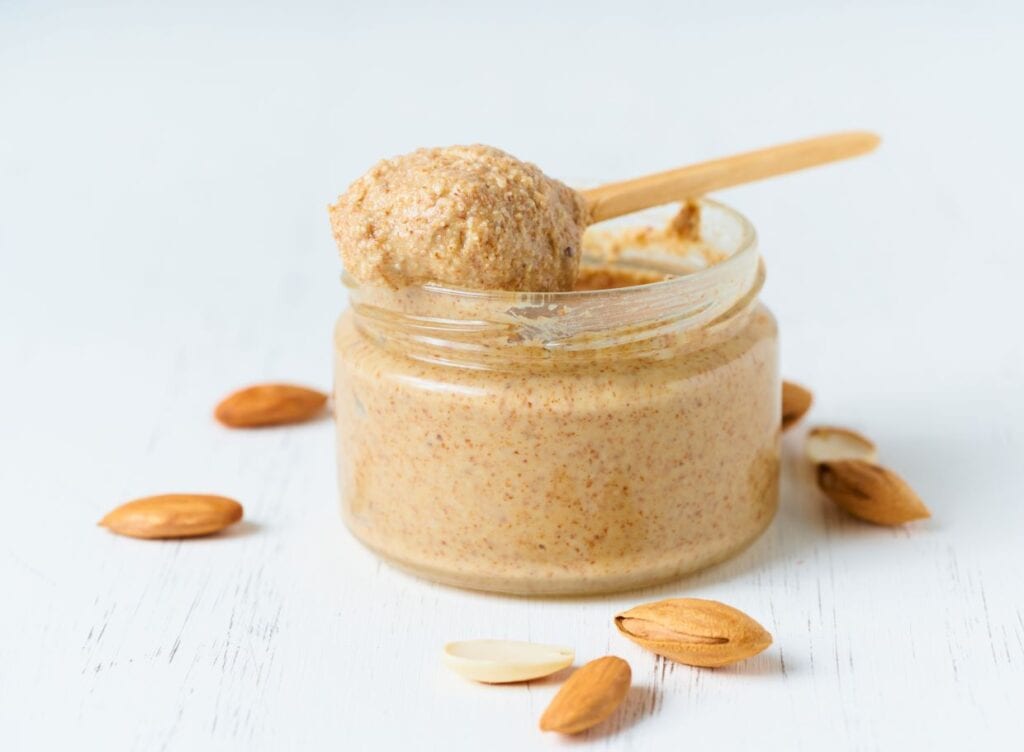 Jar of almond butter with a wooden spoon on a white surface, surrounded by almonds.