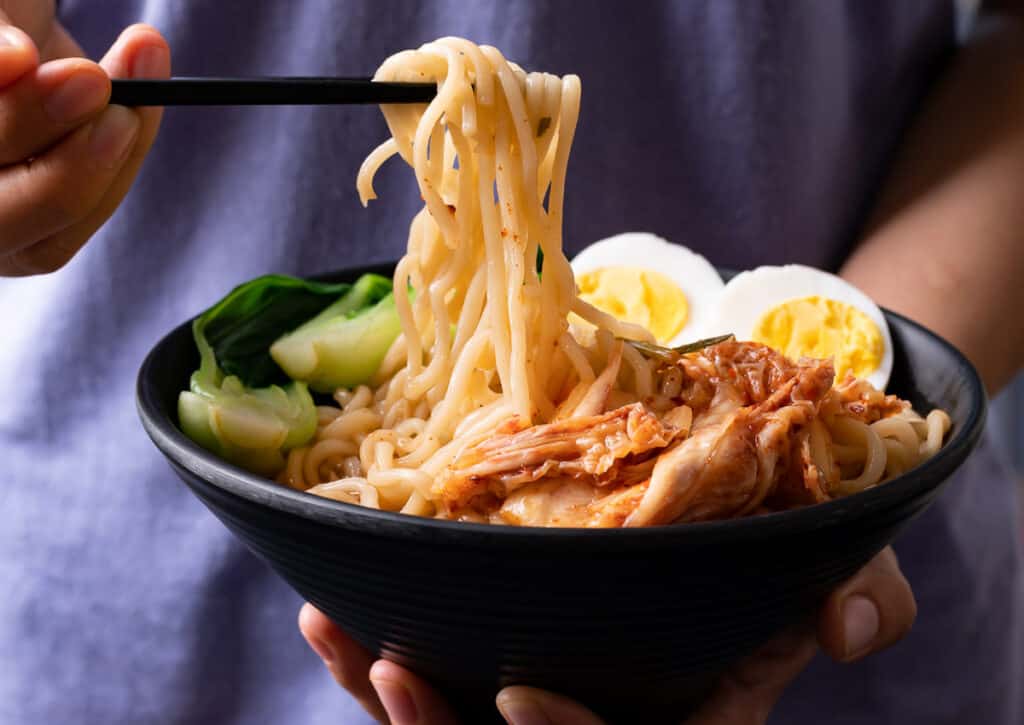 A person holding a bowl of Asian noodles with chicken and eggs.