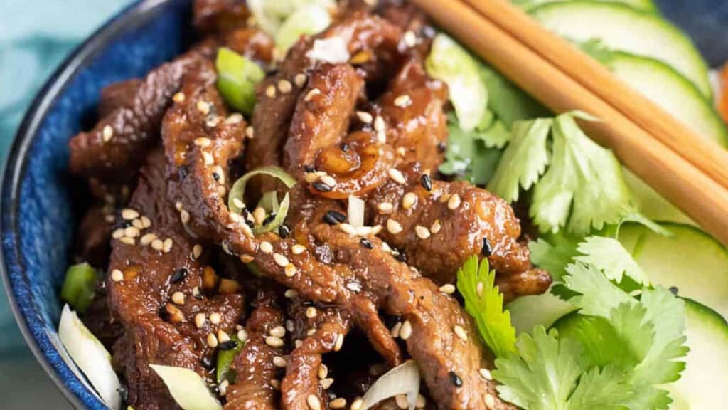 A bowl of asian-style sesame beef with cucumber slices and cilantro, accompanied by chopsticks.