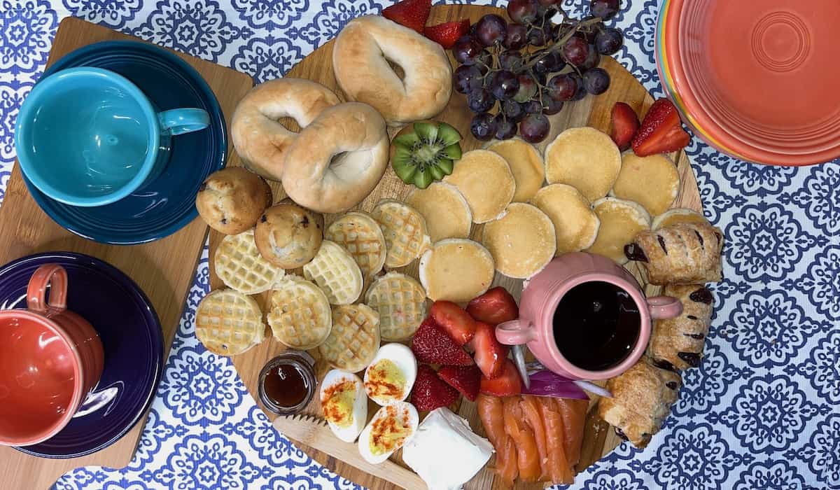 A spread of assorted breakfast foods including bagels, pastries, fruit, and coffee on a patterned tablecloth.