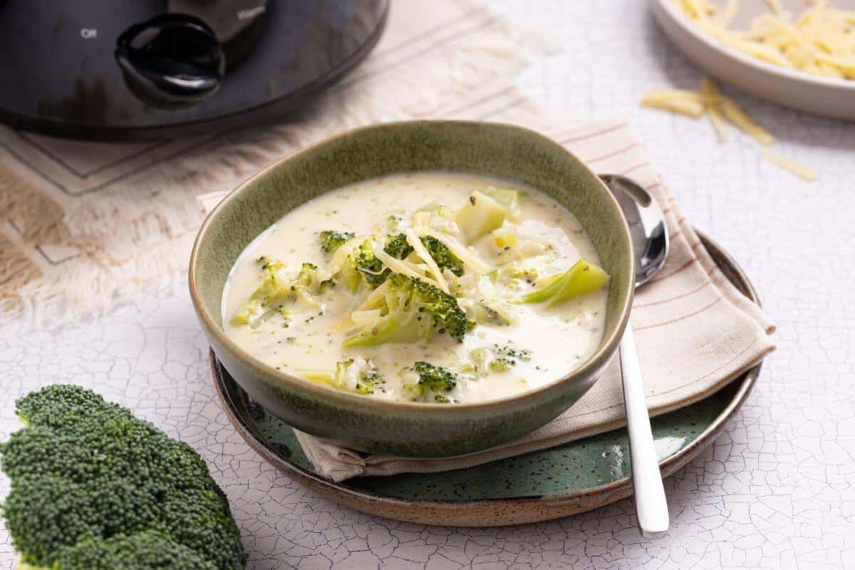 A bowl of creamy broccoli cheese soup beside fresh broccoli and shredded cheese, with a slow cooker in the background.