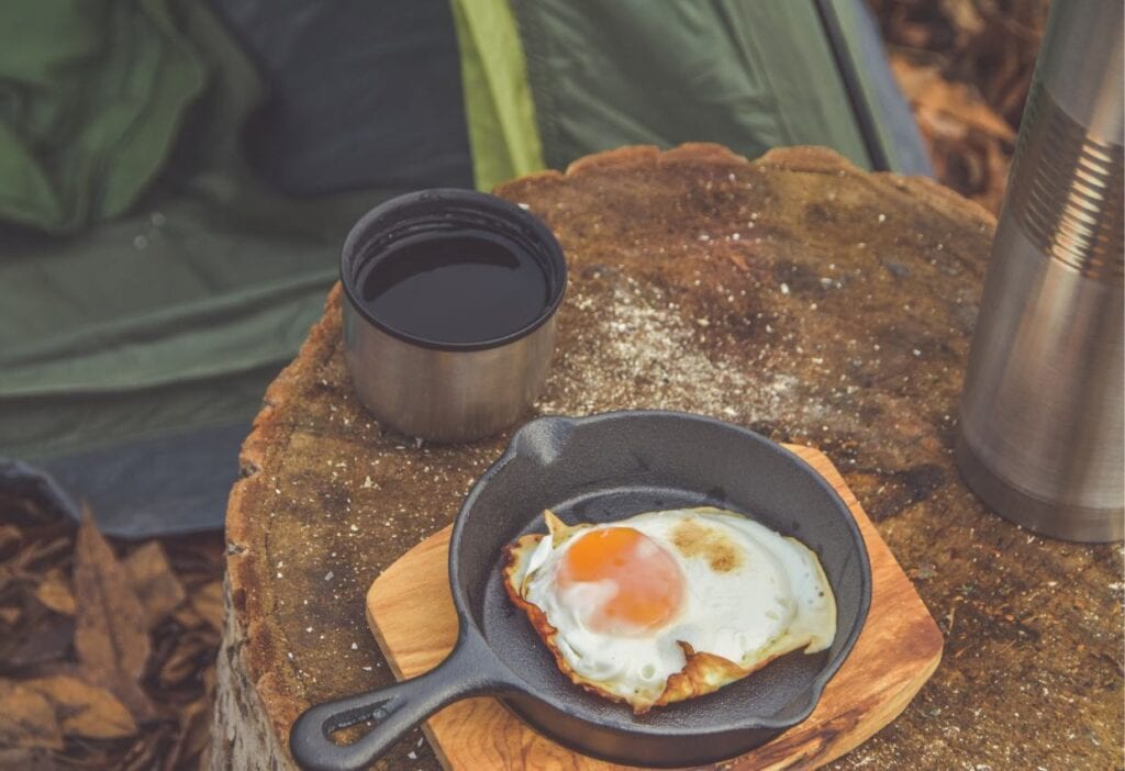 A camping food ideas breakfast scene with a fried egg on a cast iron skillet and a metal cup next to a thermos on a wooden stump.