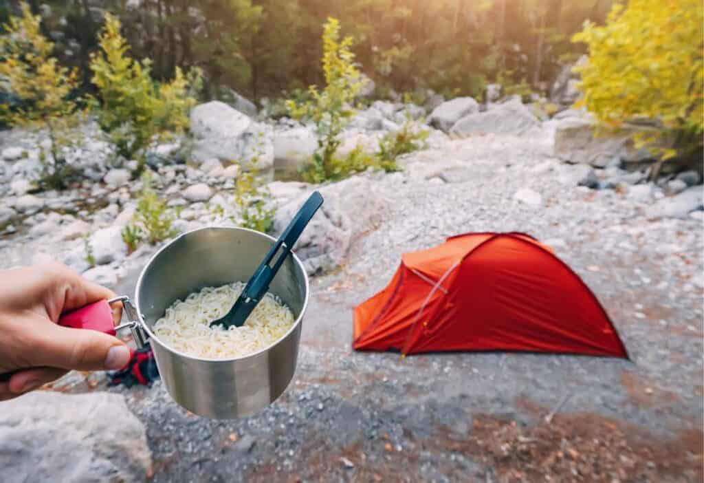 Person holding a pot of camping food ideas (noodles) with a camping tent in the background.