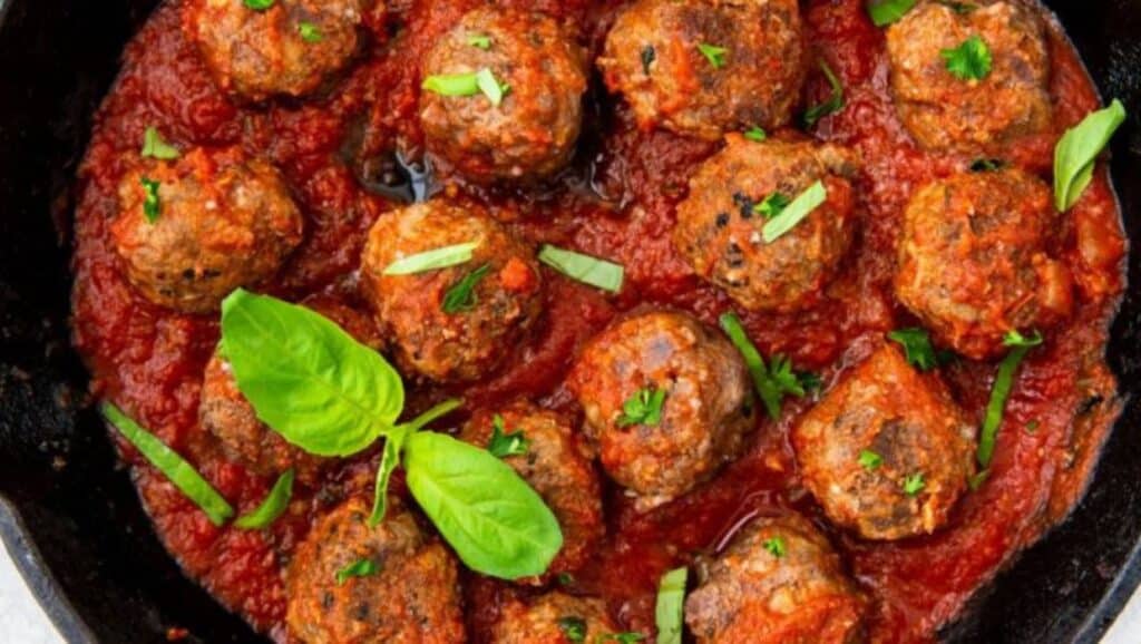 A skillet of meatballs simmering in tomato sauce garnished with fresh herbs.
