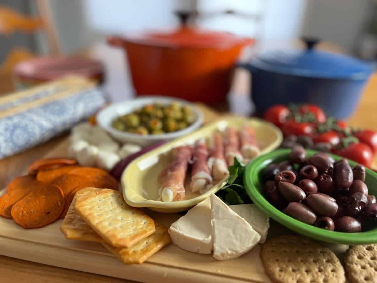 A selection of appetizers on a wooden board, featuring sliced meats, cheese, olives, tomatoes, and crackers with a colorful cookware in the background.