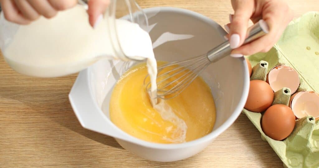 Pouring milk into a bowl of beaten eggs while whisking.