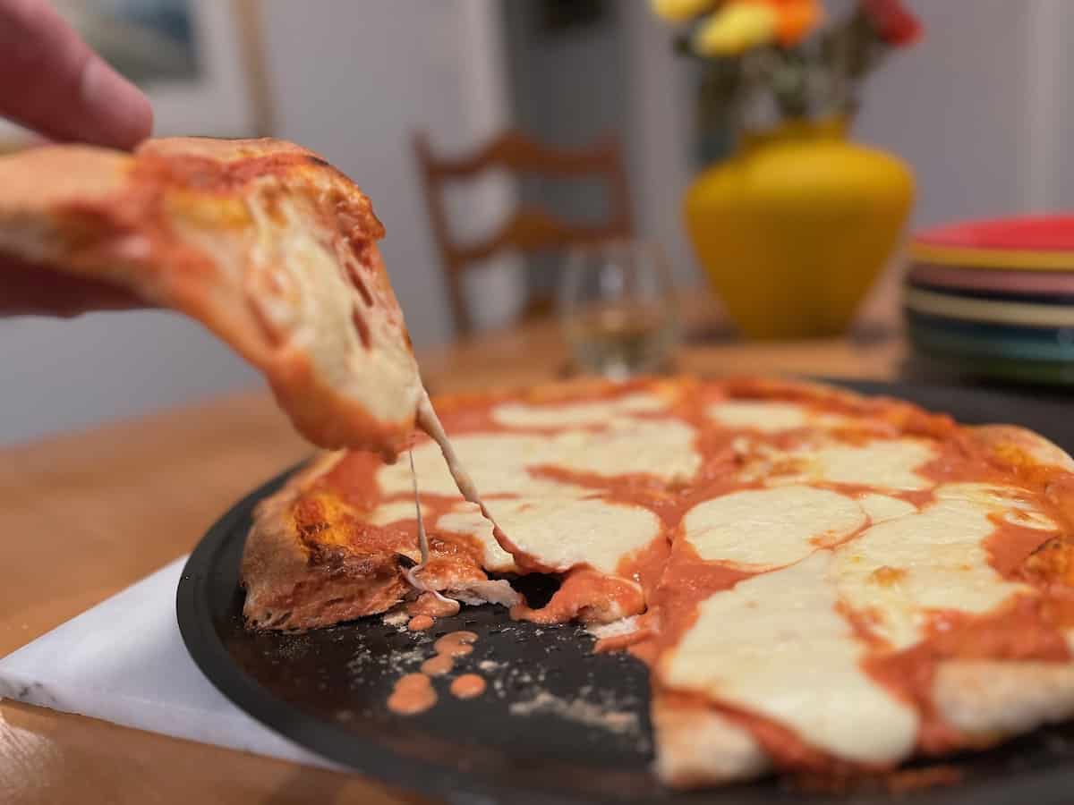 A hand holding a slice of freshly baked pizza with cheese stretching between the slice and the rest of the pizza.