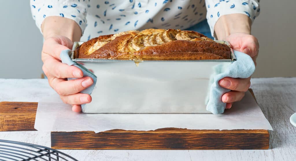 Person holding a freshly baked loaf of bread in a metal baking pan, showing how to store homemade bread properly.