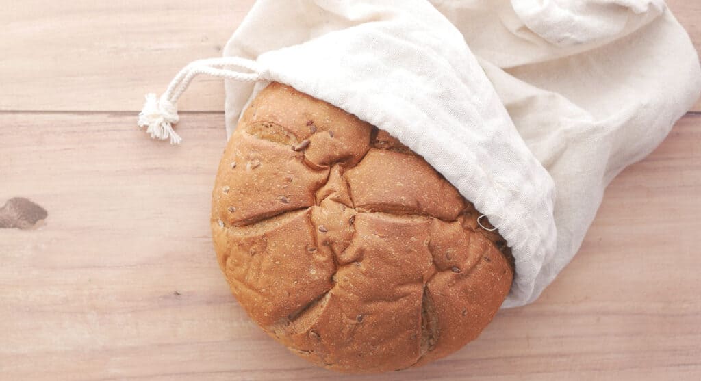 A loaf of bread partially wrapped in a cloth bag on a wooden surface demonstrates how to store homemade bread.