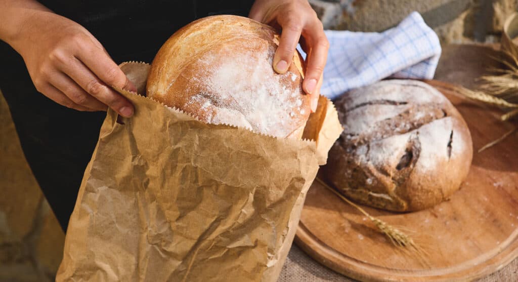 Person placing a freshly baked loaf of bread into a brown paper bag, demonstrating how to store homemade bread, with another loaf on a wooden board in the background.