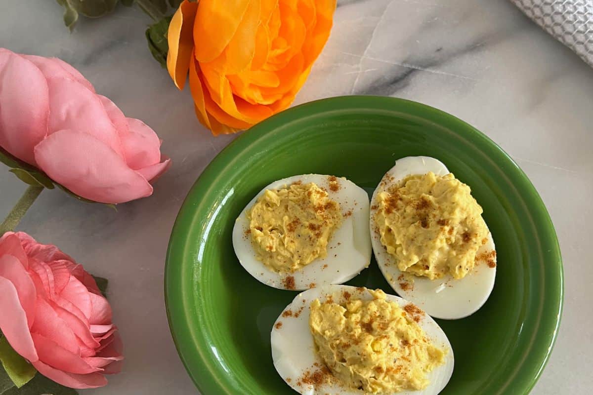 Four deviled egg halves on a green plate, garnished with paprika, accompanied by pink and orange artificial flowers.