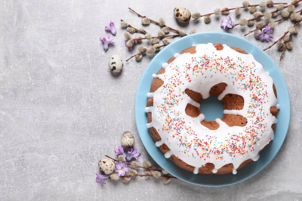 Easter-themed bundt cake with white icing and sprinkles on a blue plate, accompanied by quail eggs, pussy willow branches, and purple flowers on a textured grey background.