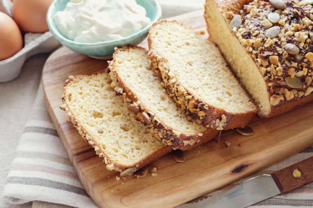 Slices of nut-topped low-carb bread with a side of cream and eggs on a wooden cutting board.
