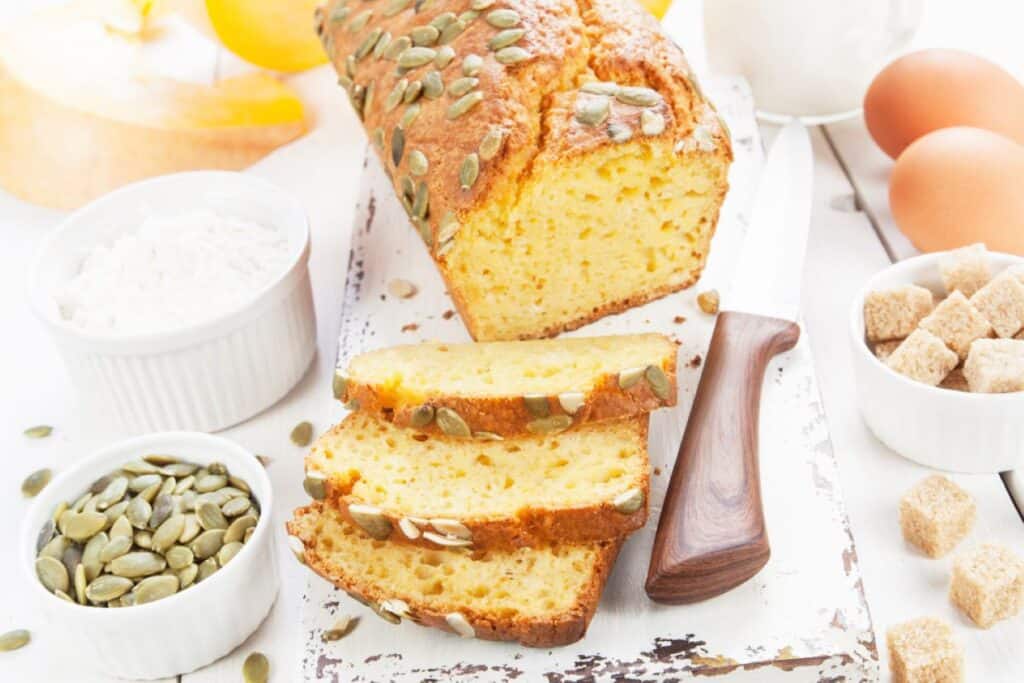 A freshly baked loaf of low-carb bread with slices, accompanied by ingredients like eggs, flour, sugar cubes, and pumpkin seeds on a white wooden background.