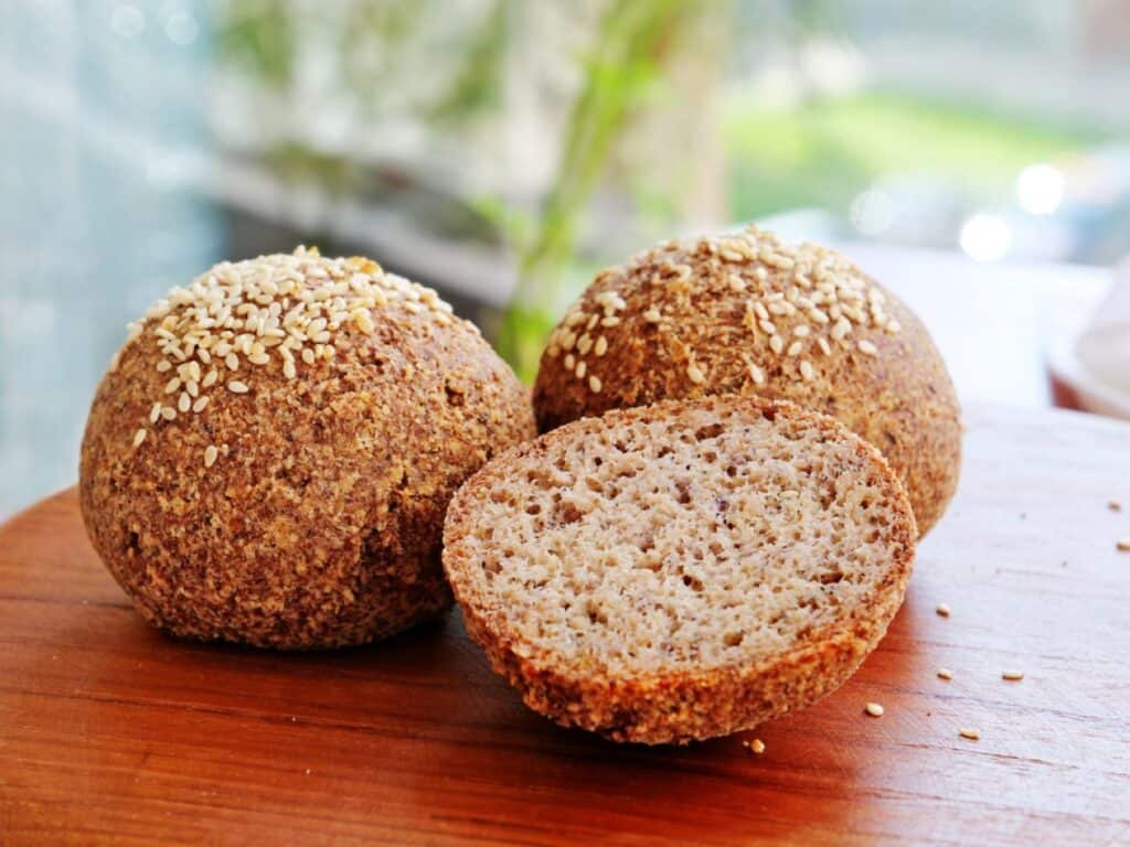 Three low-carb-bread sesame-seeded whole grain rolls on a wooden board, one sliced in half to show the texture.