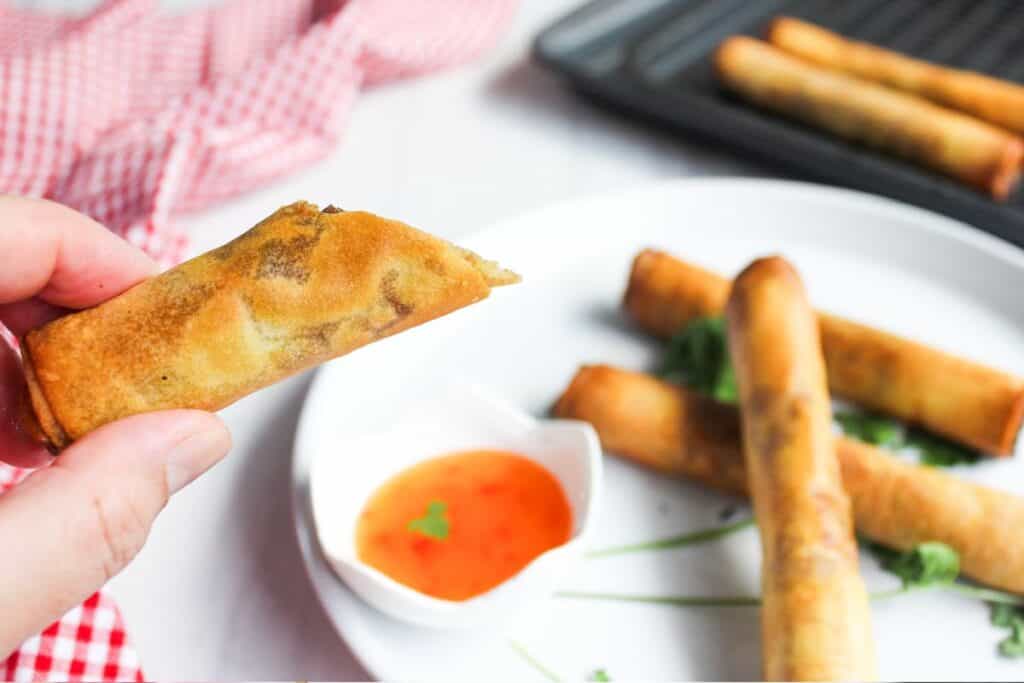 A person holding a spring roll with a side of dipping sauce and more spring rolls on a plate in the background.