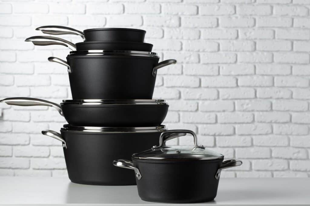 A stack of non-toxic black pots and pans with lids, against a white brick wall background.