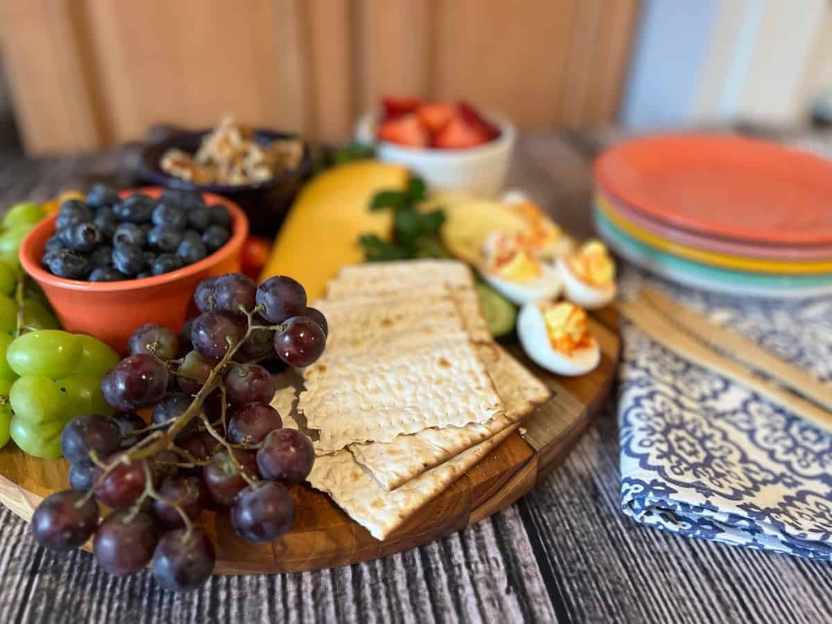 A spread of various fruits, crackers, and snacks on a wooden board with plates and utensils on the side.
