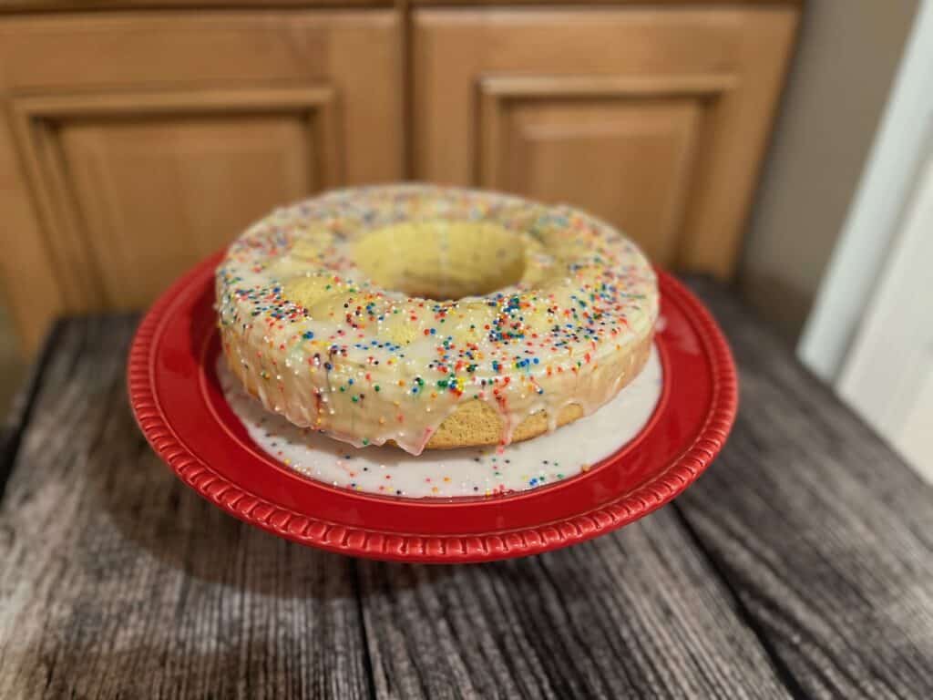 A bundt cake with white frosting and colorful sprinkles on a red plate, placed on a wooden table.