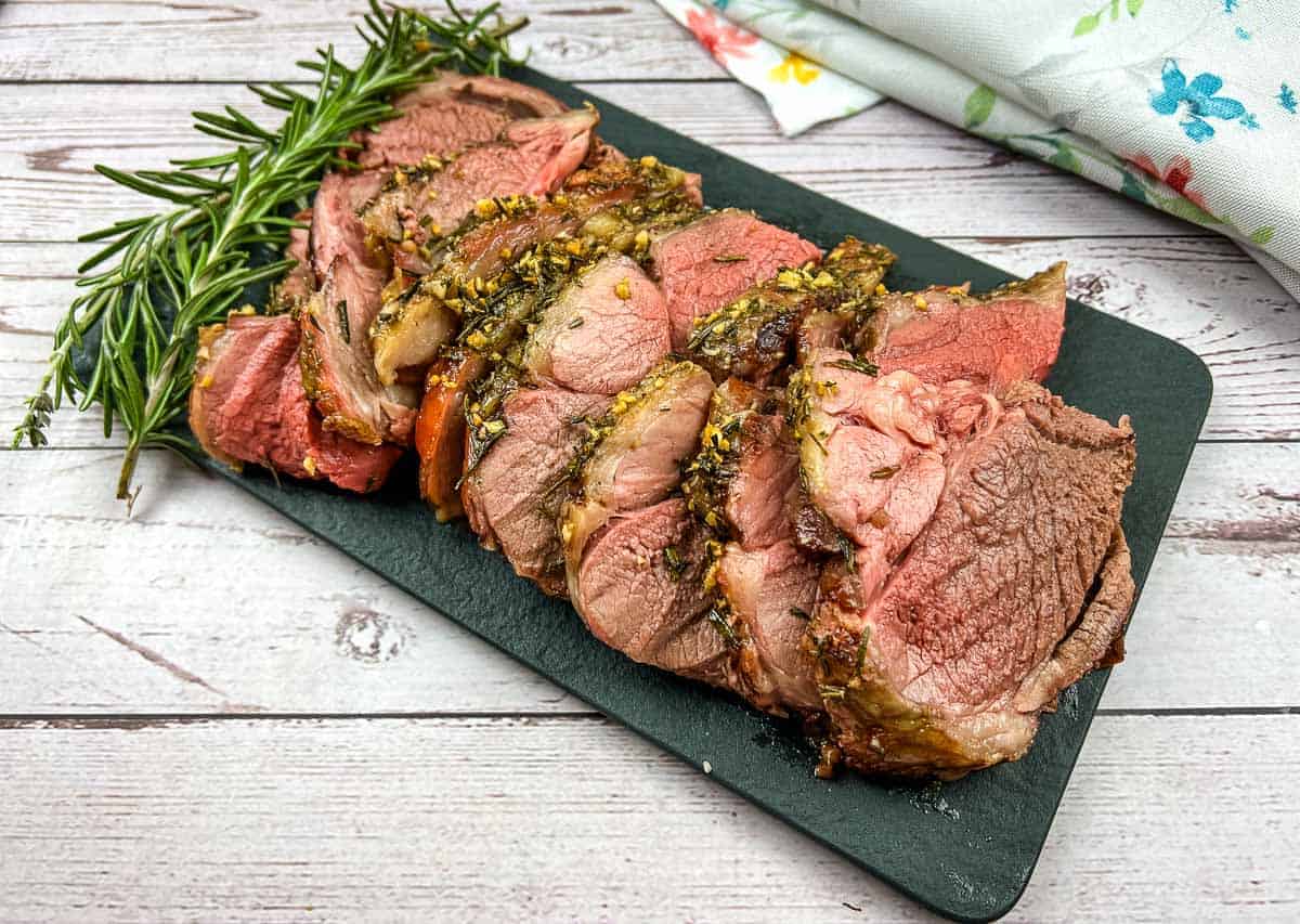 Sliced roasted leg of lamb with a herb crust on a black serving board with a sprig of rosemary on the side.