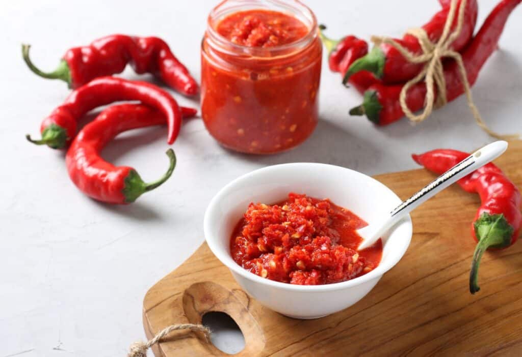 Homemade spicy red chili paste in a bowl with fresh chili peppers and a jar of paste on a kitchen countertop.