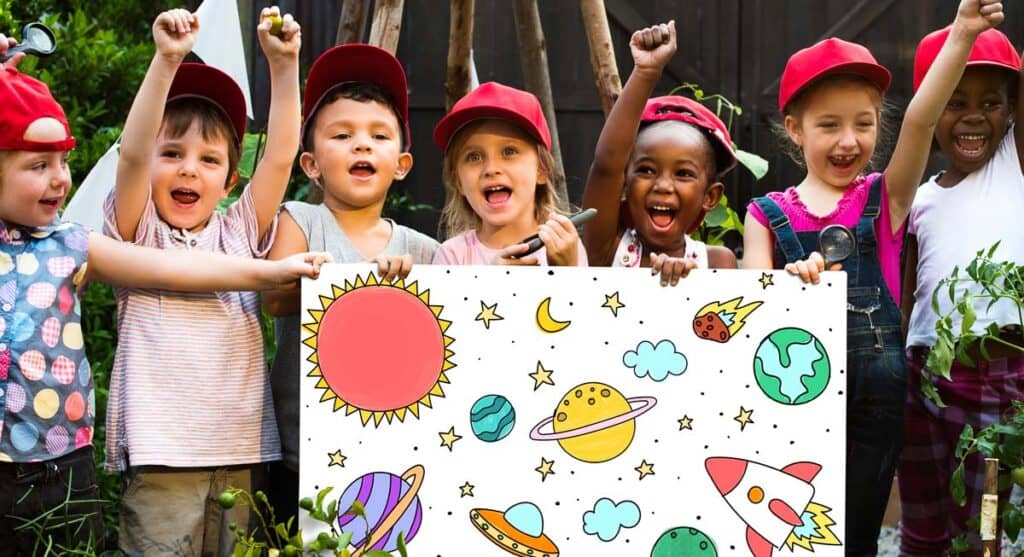 A group of joyful children wearing red caps holding up a colorful drawing of space-themed illustrations, such as the sun, planets, stars, and a rocket, outdoors.