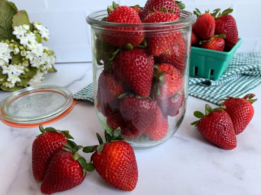 Glass jar filled with fresh strawberries on a kitchen counter, with more berries and a green basket in the background.