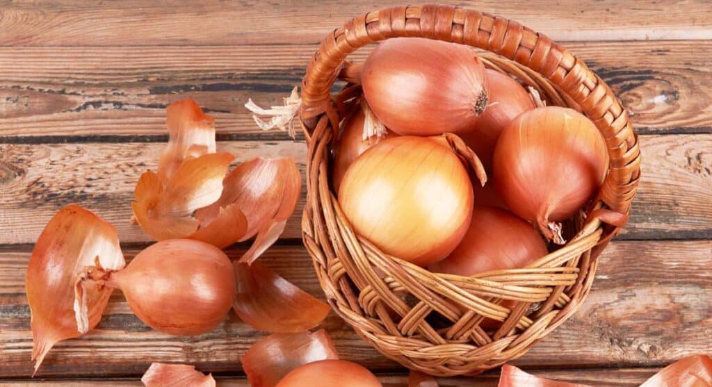 A basket full of onions on a wooden table with some onion skins scattered around.