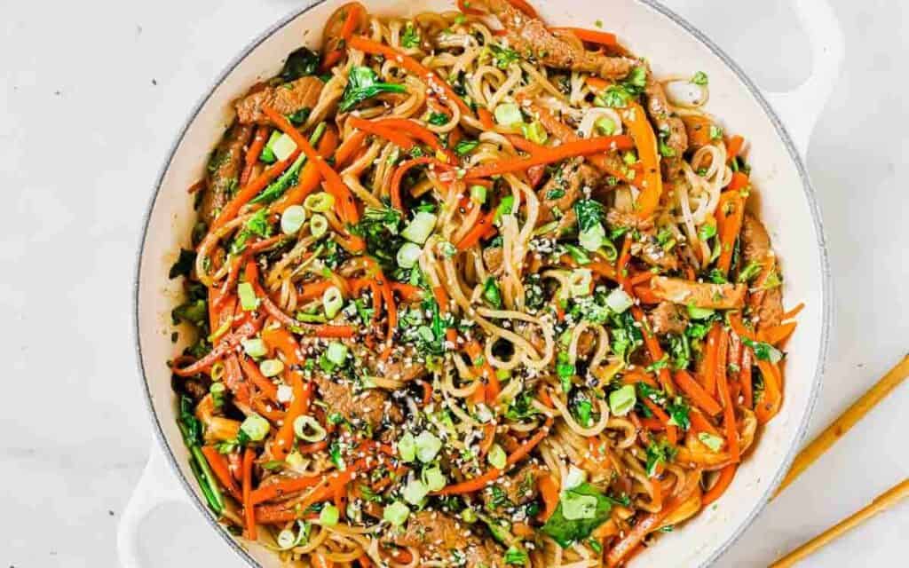 A stir-fry dish with noodles, vegetables, and sesame seeds served in a white pan.