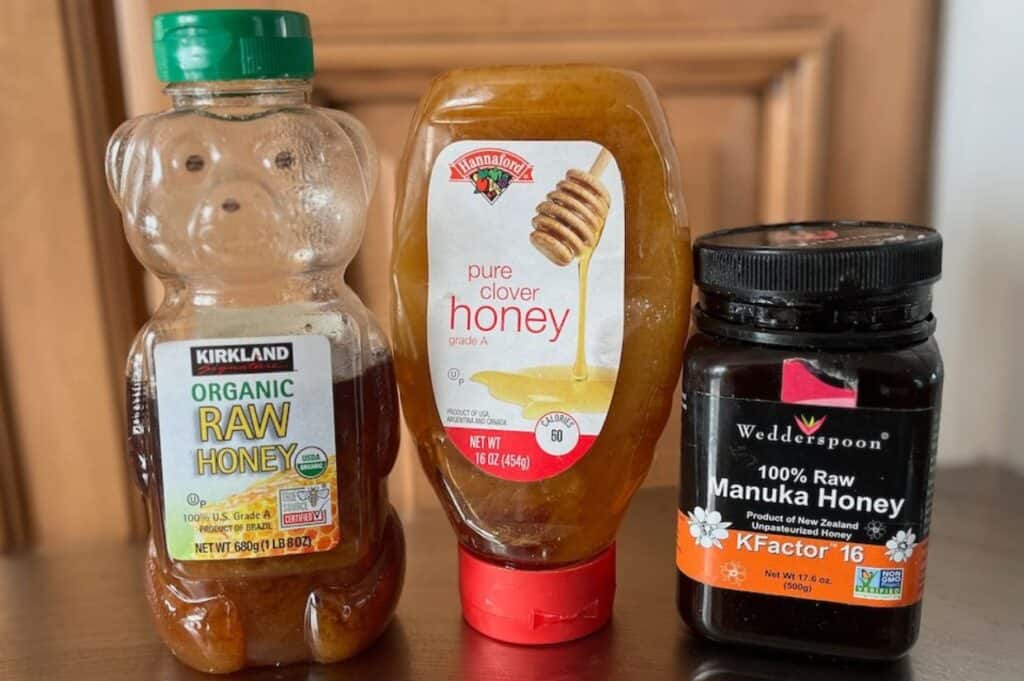 Three varieties of honey, including organic, pure grade a, and manuka ready to decrystallize, in different packaging on a wooden surface.