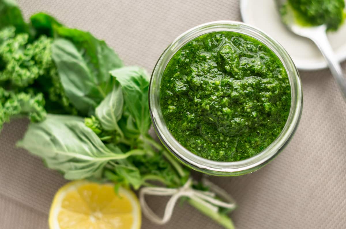 Homemade basil pesto in a glass jar surrounded by fresh basil leaves and a slice of lemon.