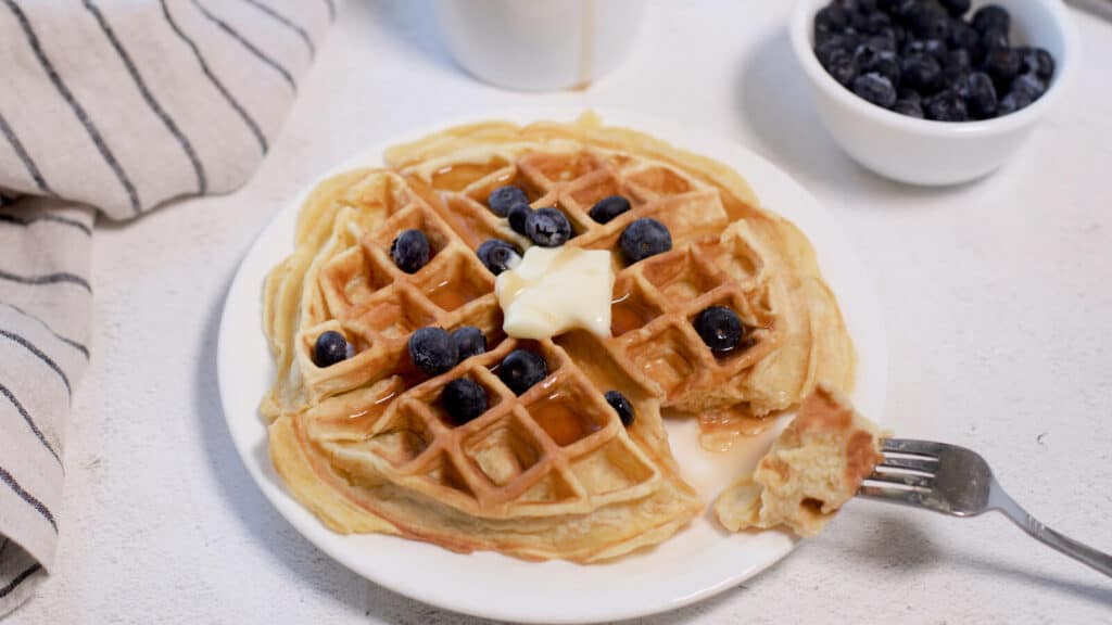 Golden waffle topped with butter and blueberries, served with maple syrup, on a white plate with a side of blueberries in a bowl.
