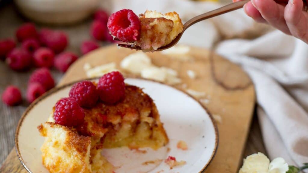A person holding a spoonful of white chocolate and raspberry bread pudding, with more of the dessert and fresh raspberries on the plate in the background.