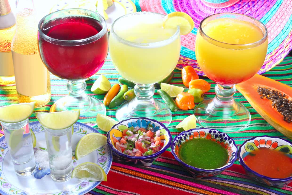 Colorful mexican drinks and appetizers on a vibrant tablecloth, including margaritas, tequila shots, and small bowls of salsa and ceviche.