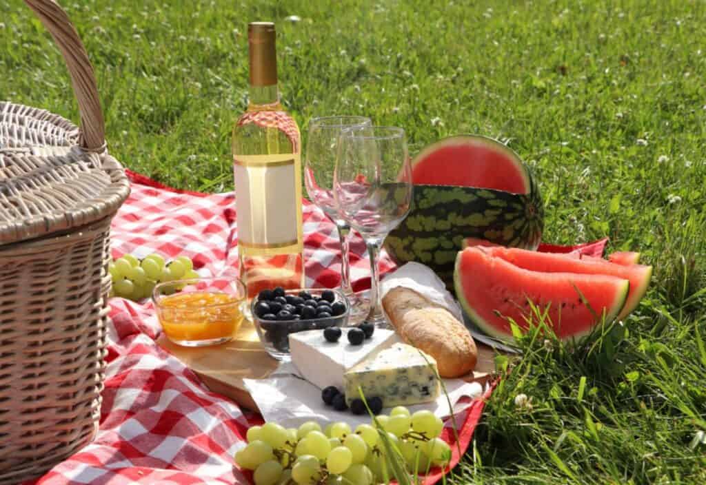 A gourmet picnic setup on grass featuring a bottle of wine, two glasses, a watermelon, grapes, blueberries, cheese, and bread beside a wicker basket.
