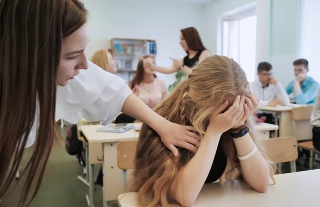 A teacher comforting a distressed student in a classroom while other students engage in conversation in the background.