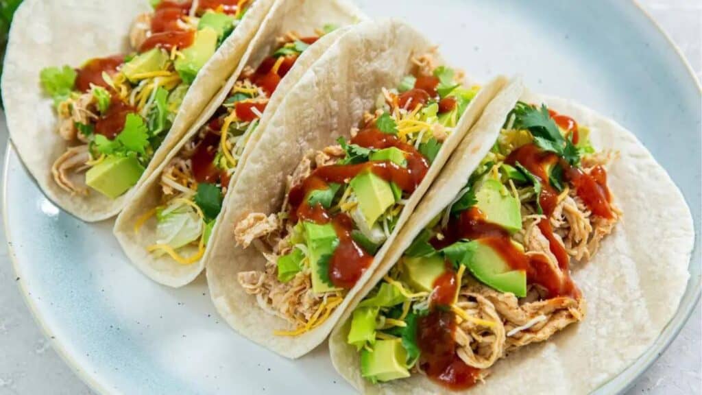 Three chicken tacos with avocado slices, shredded cheese, cilantro, and salsa on a blue plate.