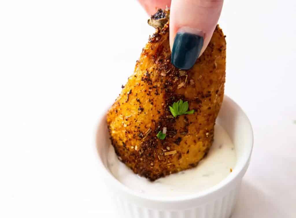A hand dipping a seasoned chicken wing into a small bowl of white sauce.